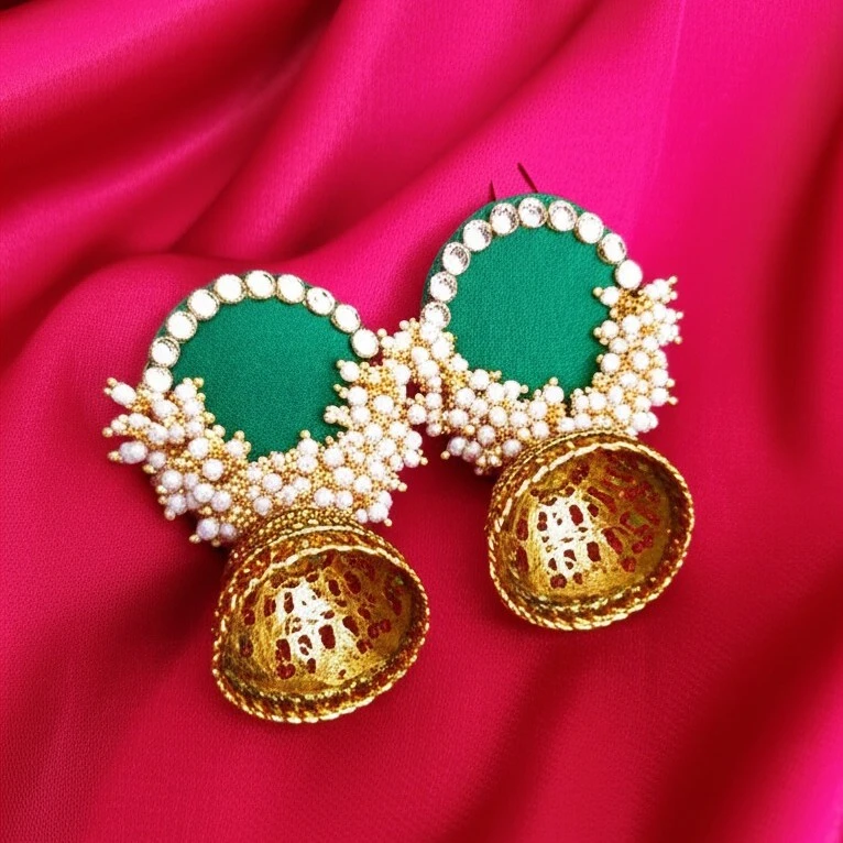 A pair of handcrafted emerald green fabric jhumka earrings with a pearl border and an antique gold filigree bell, displayed on a red background