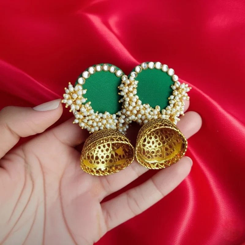 A pair of handcrafted emerald green fabric jhumka earrings with a pearl border and an antique gold filigree bell, displayed on a red background.