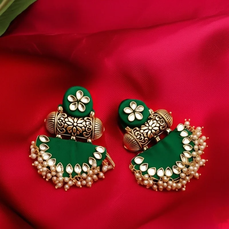 A pair of emerald green fabric chandbali earrings heavily embellished with kundan stones and a pearl border, set against a red silk background.