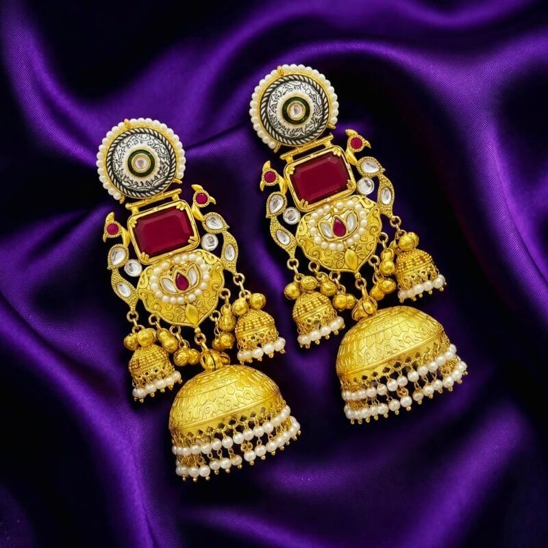 A pair of grand gold tone jhumka earrings with rectangular ruby red stones, preacock design, green enamel, and pearl detailing placed on blueish purple silk cloth
