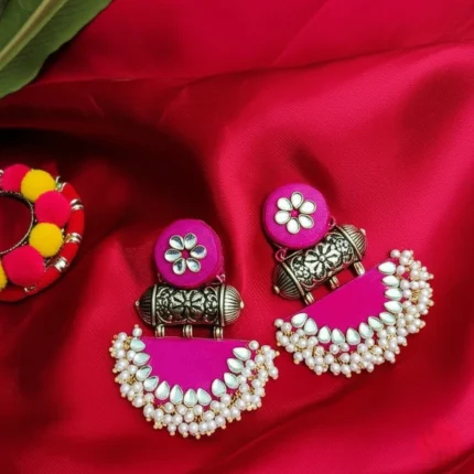 A pair of vibrant rani pink fabric chandbali earrings adorned with kundan stones and a delicate pearl border, placed on a red-colored background.
