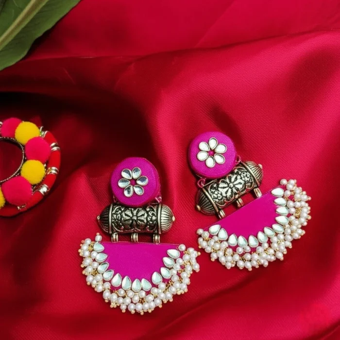 A pair of vibrant rani pink fabric chandbali earrings adorned with kundan stones and a delicate pearl border, placed on a red-colored background.