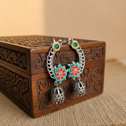 A pair of silver toned Kundan flower earcuffs with pink, turquoise, and green accents, featuring jhumka drops resting near carved wooden box