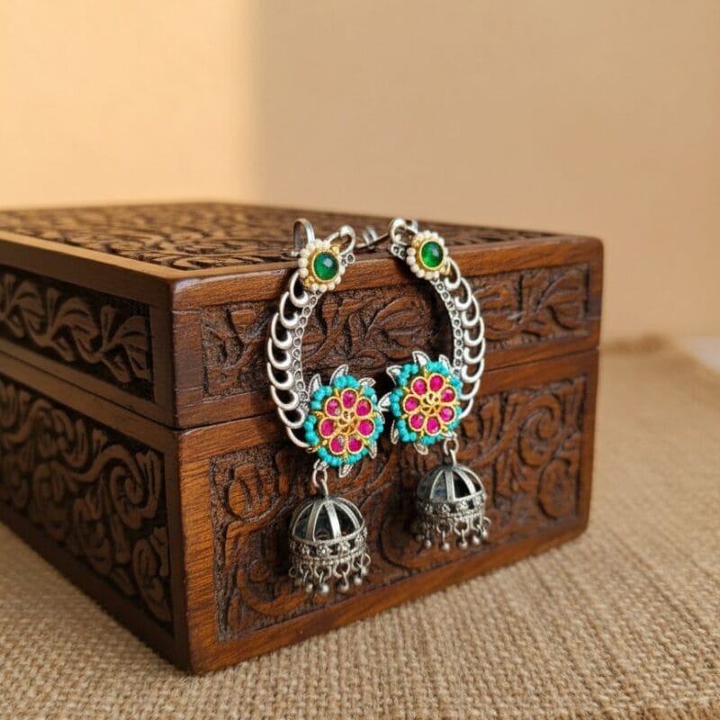 A pair of silver toned Kundan flower earcuffs with pink, turquoise, and green accents, featuring jhumka drops resting near carved wooden box