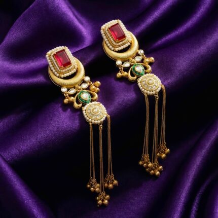 A pair of ornate golden brass Kashmiri design earrings. They feature a red rectangular stone, small pearls, green enamel details, and delicate dangling chains with small gold bells placed on blueish purple silk cloth