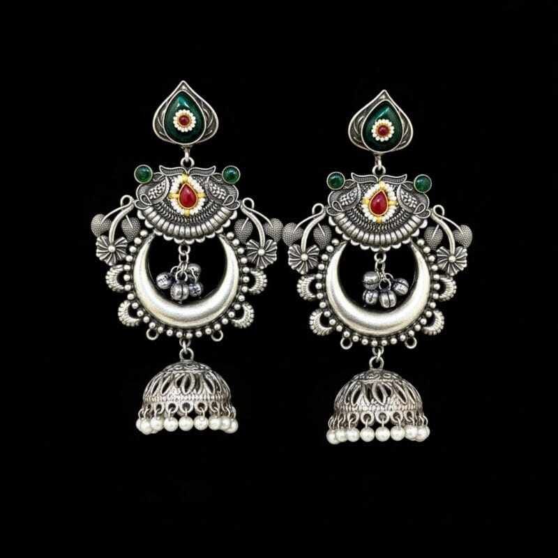 A pair of oxidized silver look alike chandbali jhumka earrings with green and red artificial stones and white pearl drops on a black background