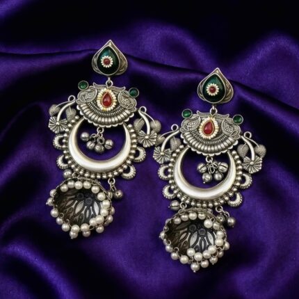 A pair of oxidized silver look alike chandbali jhumka earrings with green and red artificial stones and white pearl drops on a blueish purple silk cloth