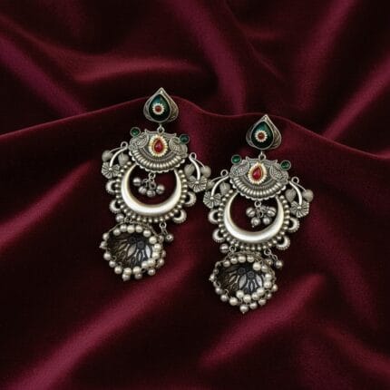 A pair of oxidized silver look alike chandbali jhumka earrings with green and red artificial stones and white pearl drops on a red silk cloth