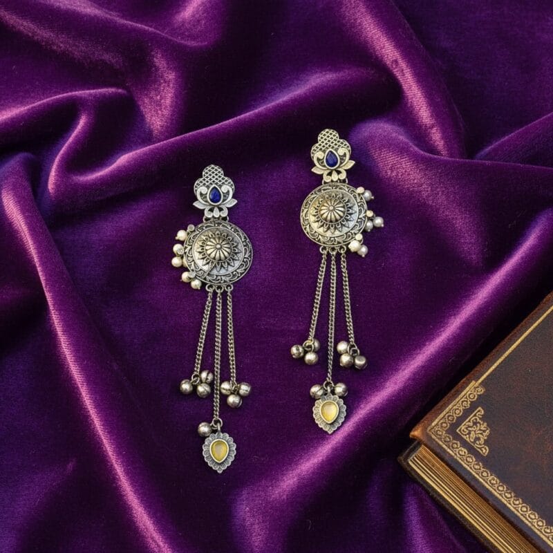 Pair of long traditional oxidized look tassel earrings with intricate patterns, blue stones at the top, and yellow heart-shaped stones at the bottom resting on purple silk cloth