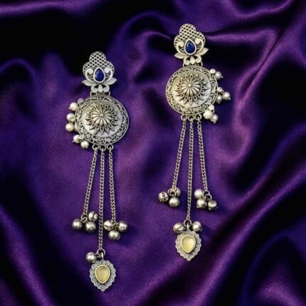 Pair of long traditional oxidized look tassel earrings with intricate patterns, blue stones at the top, and yellow heart-shaped stones at the bottom resting on purple silk cloth