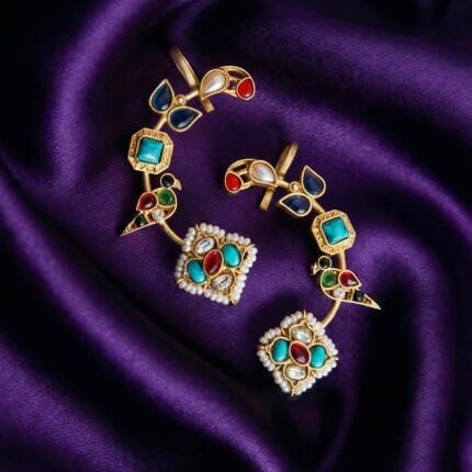A pair of colorful Rainbow Paradise Kundan earcuffs with bird designs and pearls placed on blueish purple silk cloth