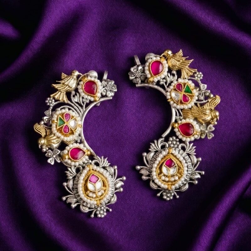 A pair of Royal Little Dove Kundan Earcuffs with silver, gold, red stones, green stones, and pearls, displayed on a bluish purple silk cloth