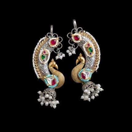 A pair of ornate royal peacock Kundan earcuffs in silver and gold tones with colorful stones, white pearls, and jhumka bells, resting on a pure black surface