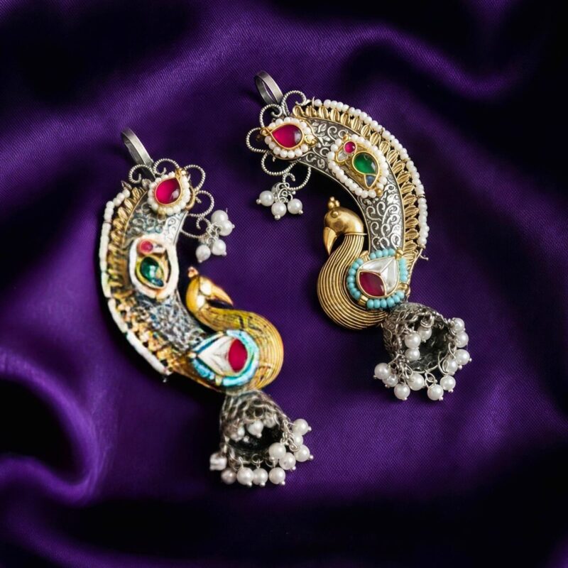 A pair of ornate royal peacock Kundan earcuffs in silver and gold tones with colorful stones, white pearls, and jhumka bells, resting on a deep purple textured fabric.