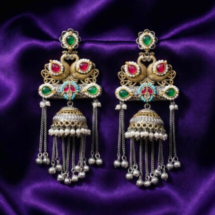 A pair of elaborate traditional Indian jhumka earrings featuring royal swan design, Kundan stones in red and green, and silver and gold tones with pearl and metal bead drops rested on a dark bluish purple silk cloth