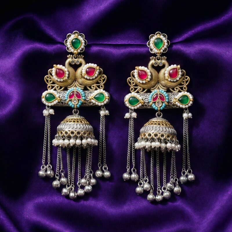A pair of elaborate traditional Indian jhumka earrings featuring royal swan design, Kundan stones in red and green, and silver and gold tones with pearl and metal bead drops rested on a dark bluish purple silk cloth
