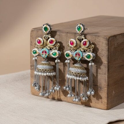 A pair of elaborate traditional Indian jhumka earrings featuring royal swan design, Kundan stones in red and green, and silver and gold tones with pearl and metal bead drops rested on a wooden block