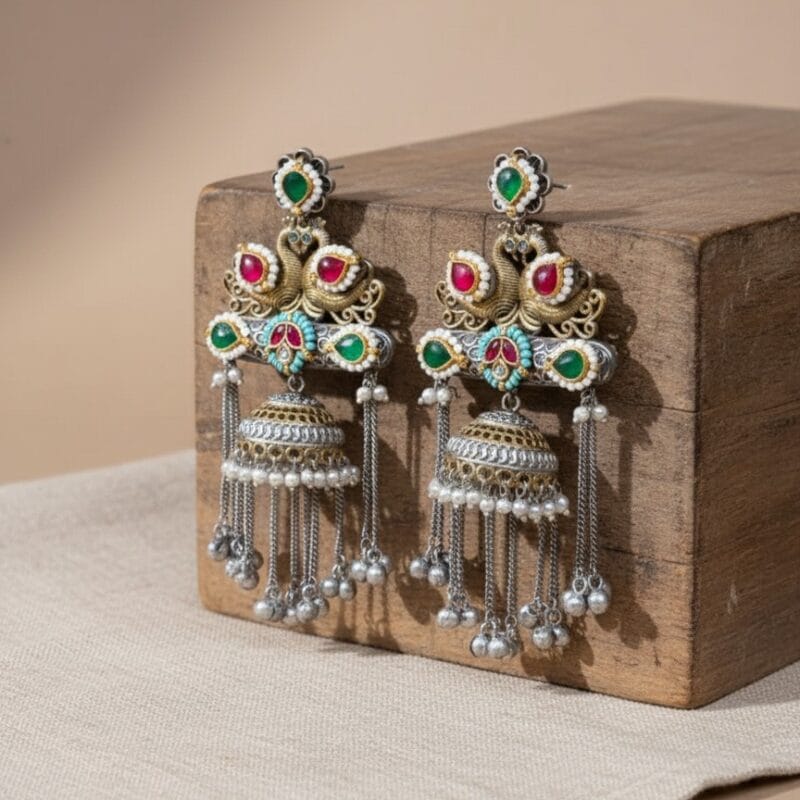A pair of elaborate traditional Indian jhumka earrings featuring royal swan design, Kundan stones in red and green, and silver and gold tones with pearl and metal bead drops rested on a wooden block