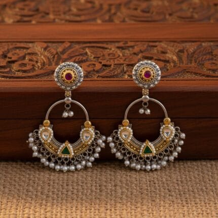A pair of traditional silver and gold Chandbali earrings with red and green stones and hanging white pearls on a carved wooden box.