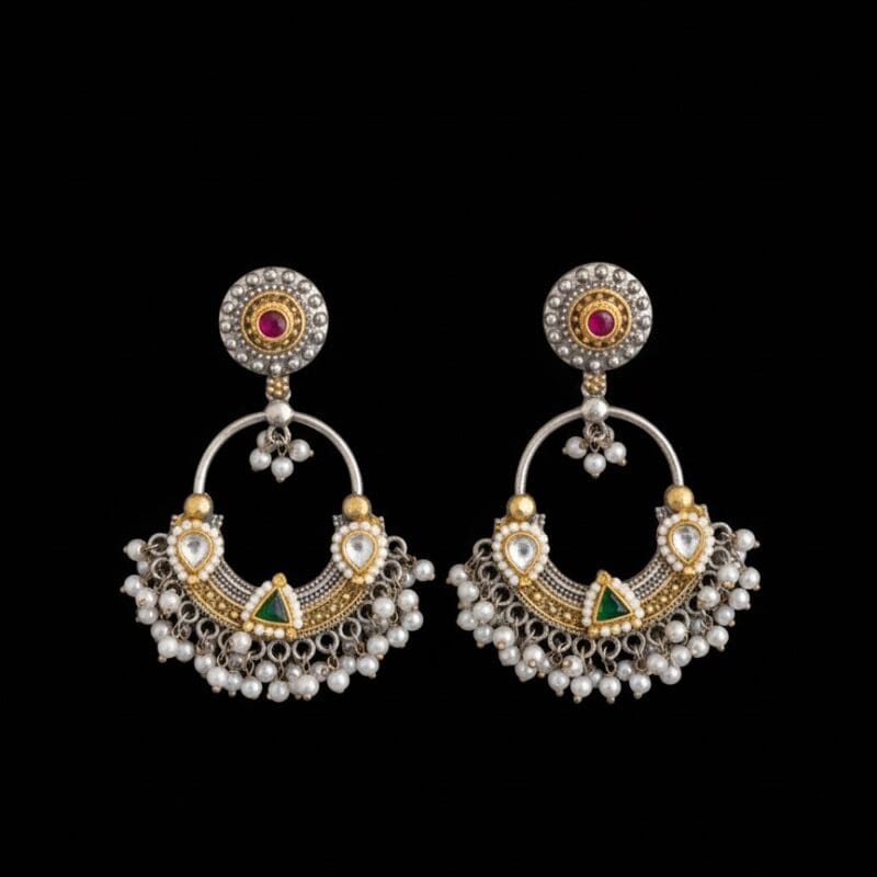 A pair of traditional silver and gold Chandbali earrings with red and green stones and hanging white pearls on a black background.