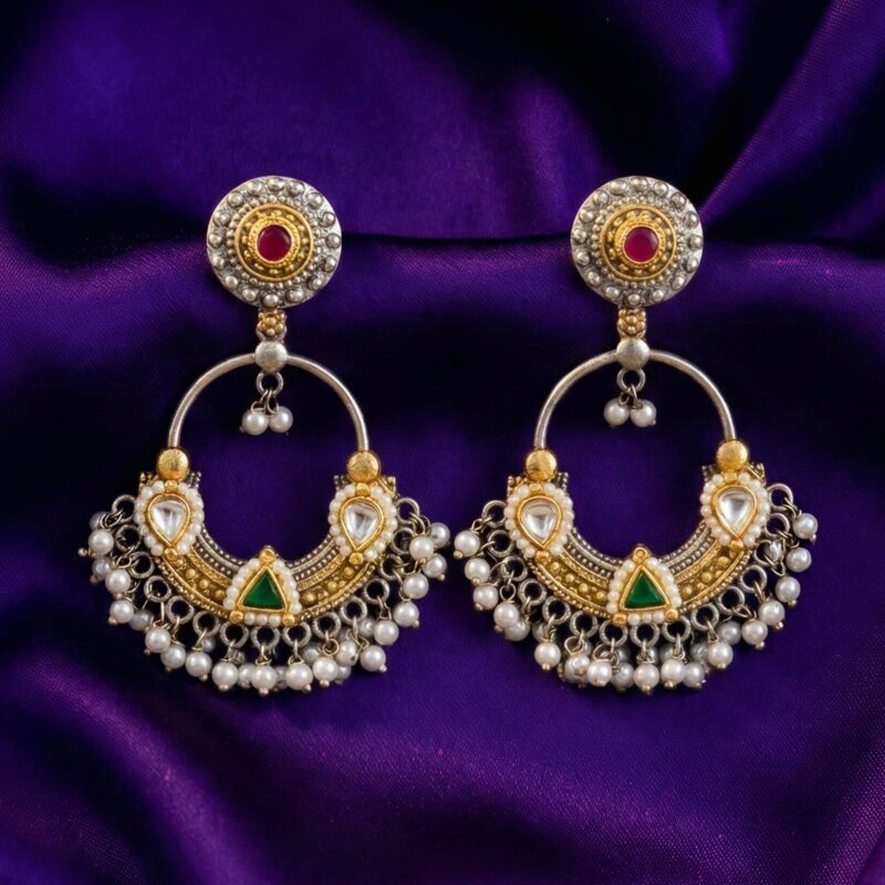 A pair of traditional silver and gold Chandbali earrings with red and green stones and hanging white pearls on a carved wooden box.
