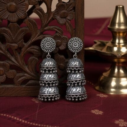 A pair of traditional three tier oxidized silver jhumka earrings with intricate floral and leaf patterns and dangling beads, resting on Traditional, Indian Aesthetic