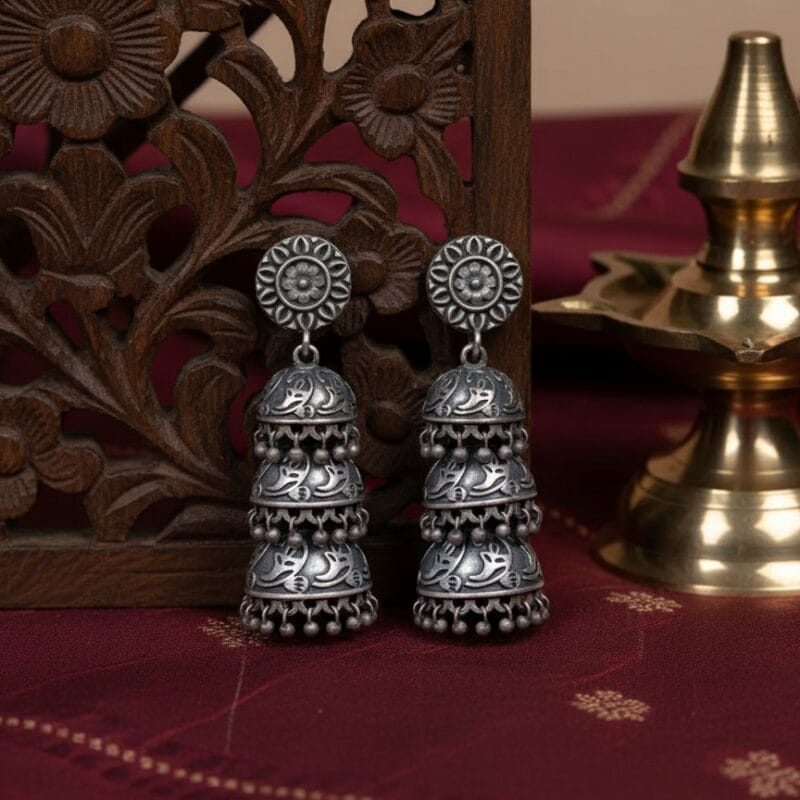 A pair of traditional three tier oxidized silver jhumka earrings with intricate floral and leaf patterns and dangling beads, resting on Traditional, Indian Aesthetic