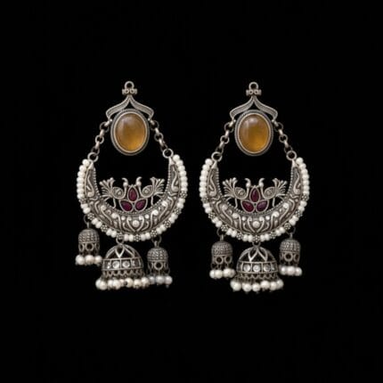 A pair of ornate oxidized silver Chandbali earrings with a yellow oval stone, ruby-red peacock designs, white pearls, and three dangling jhumkas, displayed displayed on pure black background