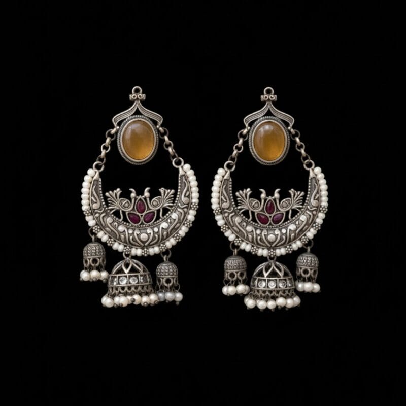 A pair of ornate oxidized silver Chandbali earrings with a yellow oval stone, ruby-red peacock designs, white pearls, and three dangling jhumkas, displayed displayed on pure black background