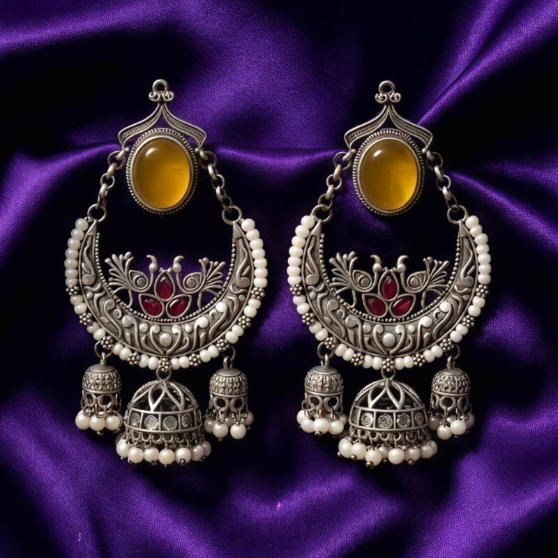 A pair of ornate oxidized silver Chandbali earrings with a yellow oval stone, ruby-red peacock designs, white pearls, and three dangling jhumkas placed on dark purple silk cloth