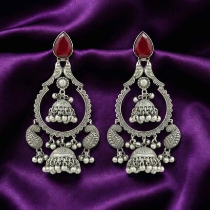 A pair of traditional oxidized silver look earrings with red teardrop stones, two birds, jhumka bells, and peacock designs on blueish purple silk cloth