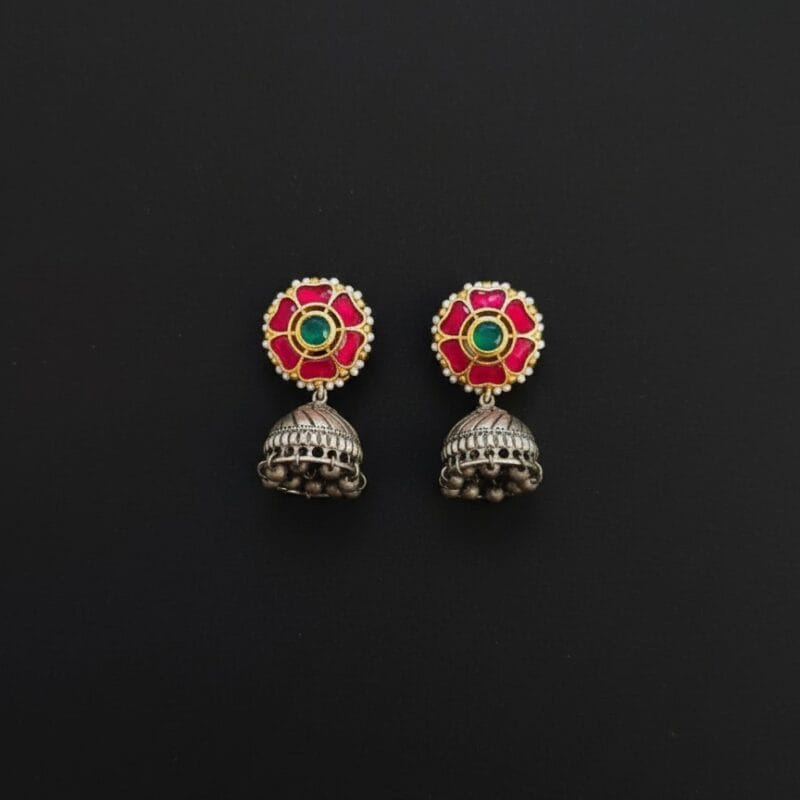 A pair of traditional Indian earrings featuring a pink flower stud with a green center stone and white pearls, connected to an oxidized silver-tone jhumka with small dangling bells placed on black background