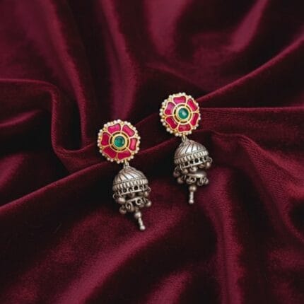A pair of traditional Indian earrings featuring a pink flower stud with a green center stone and white pearls, connected to an oxidized silver-tone jhumka with small dangling bells.