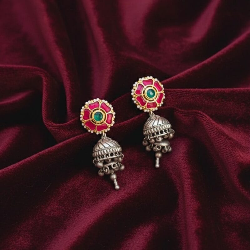 A pair of traditional Indian earrings featuring a pink flower stud with a green center stone and white pearls, connected to an oxidized silver-tone jhumka with small dangling bells.