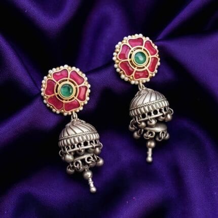 A pair of traditional Indian earrings featuring a pink flower stud with a green center stone and white pearls, connected to an oxidized silver-tone jhumka with small dangling bells.