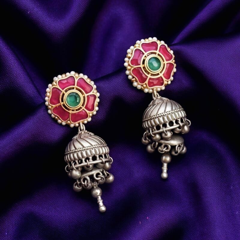 A pair of traditional Indian earrings featuring a pink flower stud with a green center stone and white pearls, connected to an oxidized silver-tone jhumka with small dangling bells.