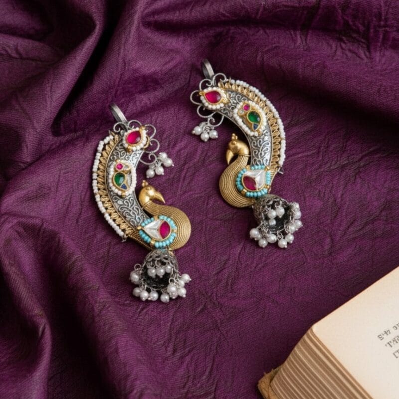 A pair of ornate royal peacock Kundan earcuffs in silver and gold tones with colorful stones, white pearls, and jhumka bells, resting on a deep purple textured fabric.