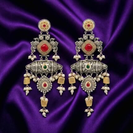 A pair of detailed traditional silver look earrings with red and green stones, white pearls, and gold accents on blueish purple silk cloth