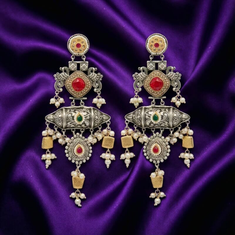 A pair of detailed traditional silver look earrings with red and green stones, white pearls, and gold accents on blueish purple silk cloth