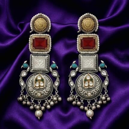 A pair of long traditional silver look earrings with square red Kundan stones, small pearls, teal accents, and silver ghungroos on blueish purple silk cloth
