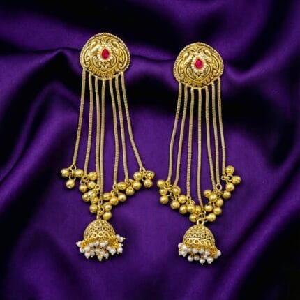 A pair of ornate golden jhumka chain earrings with a pink stone and white pearls on blueish purple silk cloth