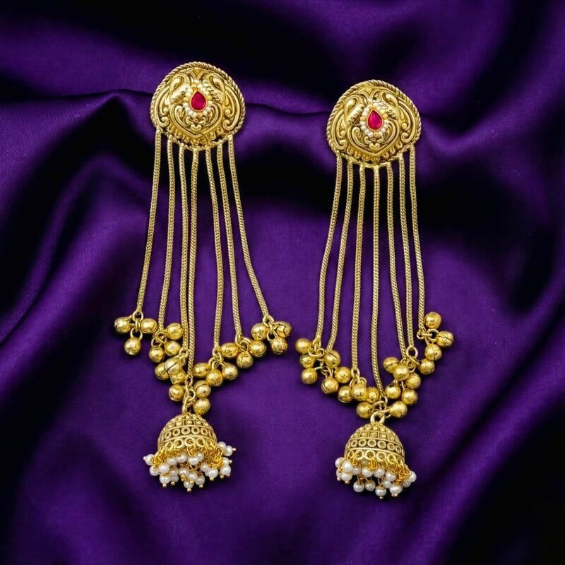 A pair of ornate golden jhumka chain earrings with a pink stone and white pearls on blueish purple silk cloth