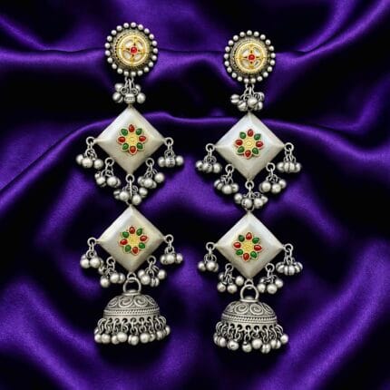 A pair of long traditional oxidized silver look jhumka earrings featuring square elements with red, green, and yellow artificial stones placed on blueish purple silk cloth