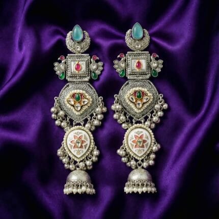 a pair of long silver plated traditional Kundan Chandbali Jhumka earrings with blue, pink, and green artificial stones placed on a blueish purple silk cloth