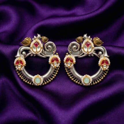 A pair of traditional Kundan brass earrings with silver and gold tones, red kundan stones, small white pearls, and a light green center stone placed on blueish purple silk cloth