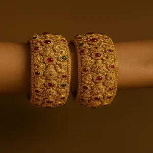 A pair of ornate gold bangles worn on a wrist. The bangles are decorated with intricate floral and circular motifs, embedded with red and green gemstones
