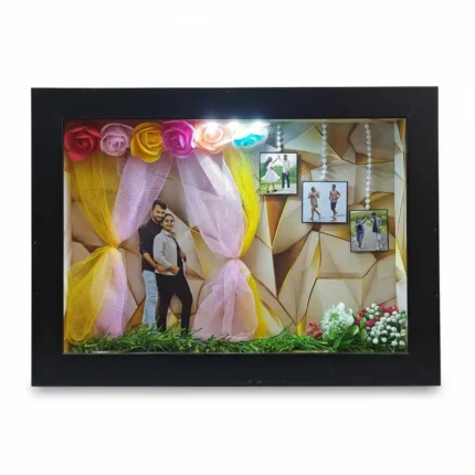 Modern black 3D shadow box frame with gold geometric background with top spotlights, couple photo cutout, hanging miniature prints.